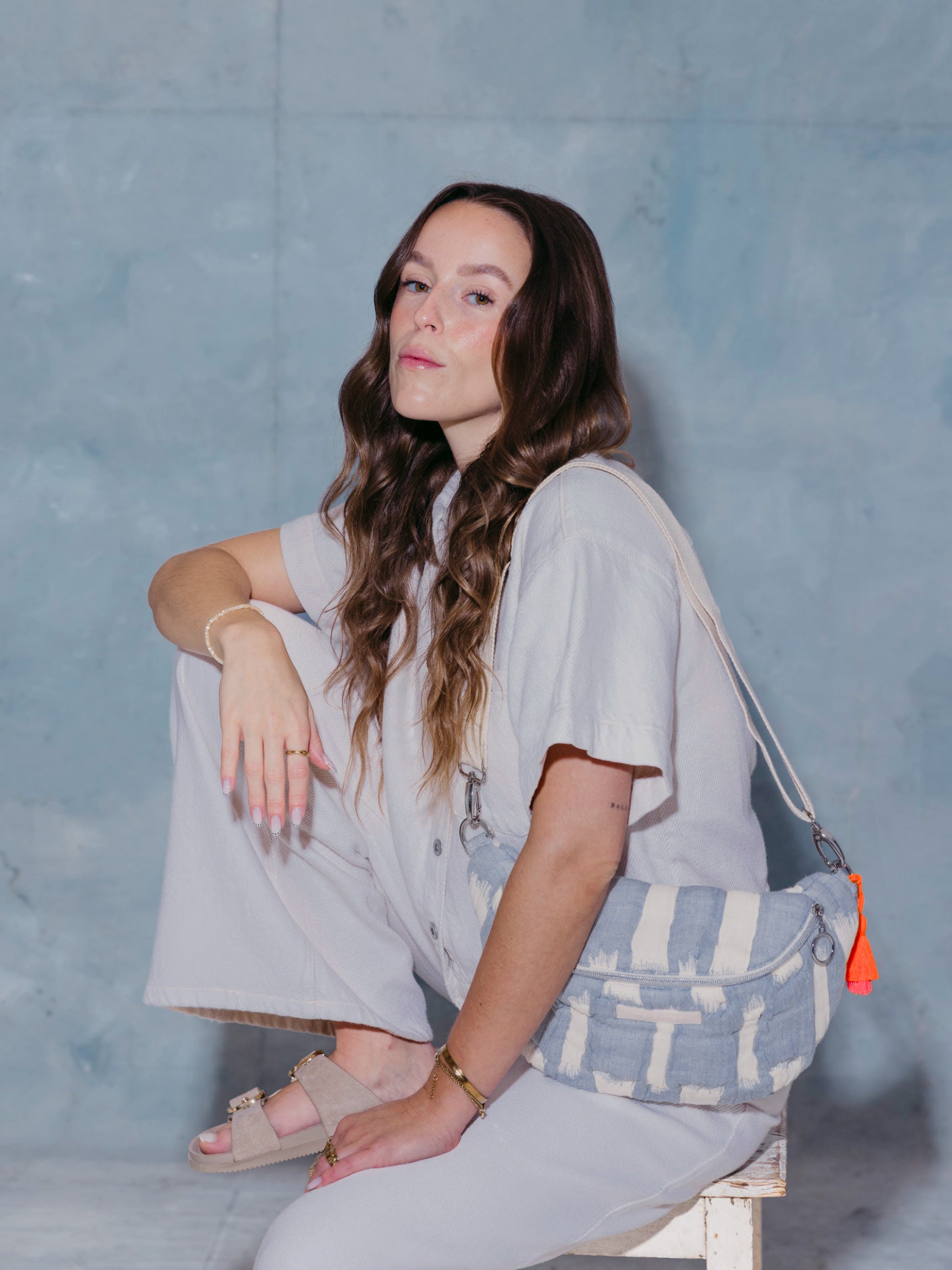 Frau sitzt mit der mara mea Midi Bauchtasche creamy skies in Hellblau-Offwhite, verstellbarem Schultergurt und neon-orange-pinker Quaste auf Hocker