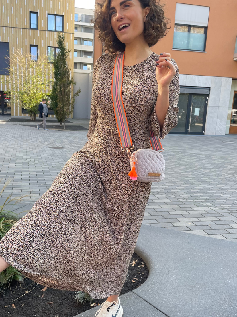 Breiter Extra-Tragegurt in knalligen Neonfarben an grauer Crossbody-Bag aus abgestepptem Samt und passender Quaste in Pink/Orange an Model mit langem Blumenkleid.