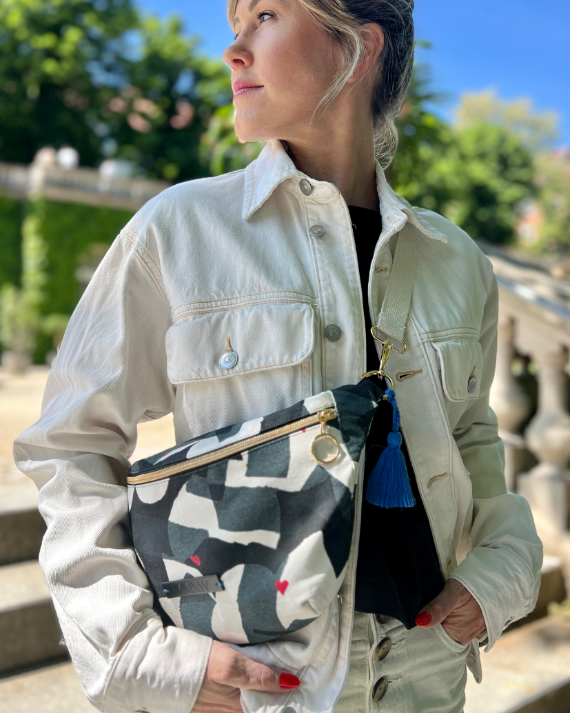 Midi Bumbag mit schwarz/weiß/grauem Print und goldenem Reißverschluss, blauer Quaste und roten aufgedruckten Herzchen Details.