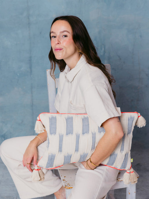 Frau sitzt mit mara mea Kissen cotton mood in Hellblau mit Offwhite-Muster, neon-oranger Paspel und Quasten