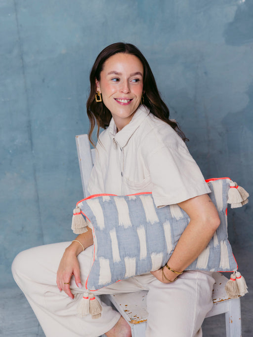 Frau sitzt mit mara mea Kissen cotton mood in Hellblau mit Offwhite-Muster, neon-oranger Paspel und Quasten
