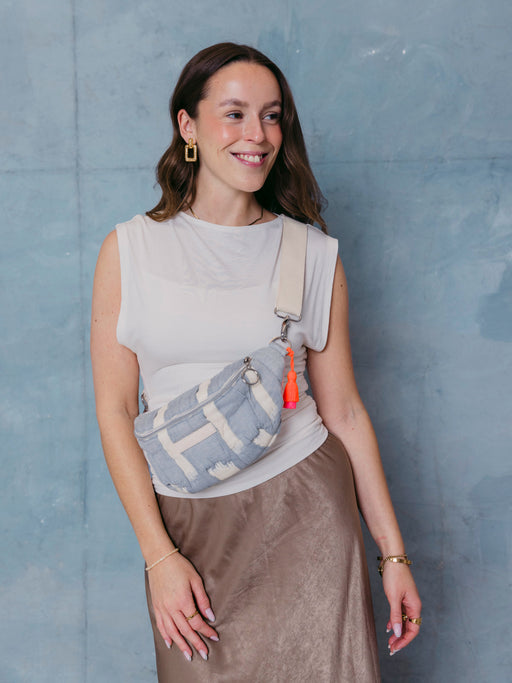 Frau trägt die mara mea Mini Bauchtasche cloud club in Hellblau-Offwhite mit breitem  verstellbarem Schultergurt, silbernem Ring-Zipper und neon-orange-pinker Quaste