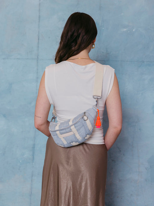 Frau trägt die mara mea Mini Bauchtasche cloud club in Hellblau-Offwhite mit verstellbarem Gurt und neon-orange-pinker Quaste auf dem Rücken