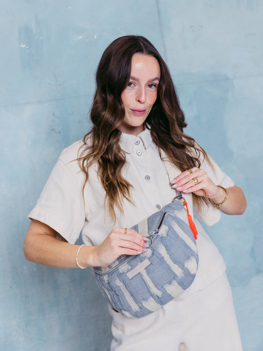 Frau öffnet die mara mea Midi Bauchtasche creamy skies in Hellblau-Offwhite mit Ring-Zipper, Markenlabel und neon-orange-pinker Quaste