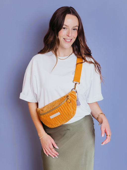 Frau trägt die orangefarbene mara mea Mini Bauchtasche Come Dancing Peach aus Cord quer über der Hüfte vor blauem Hintergrund
