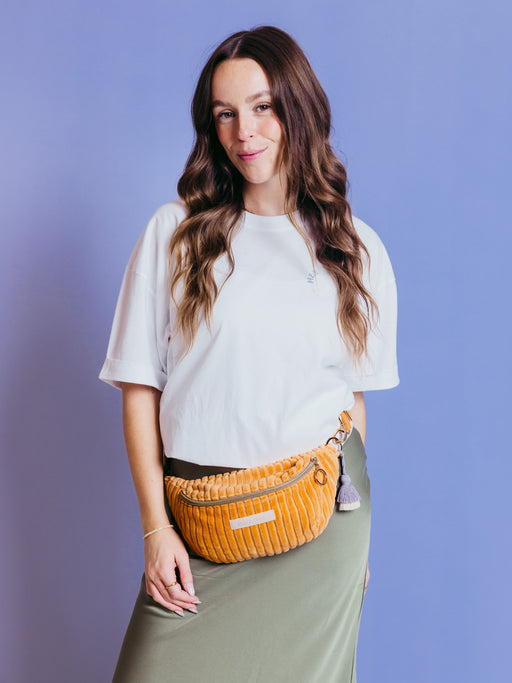 Frau trägt die orangefarbene mara mea Mini Bauchtasche Come Dancing Peach aus Cord um die Hüfte vor blauem Hintergrund