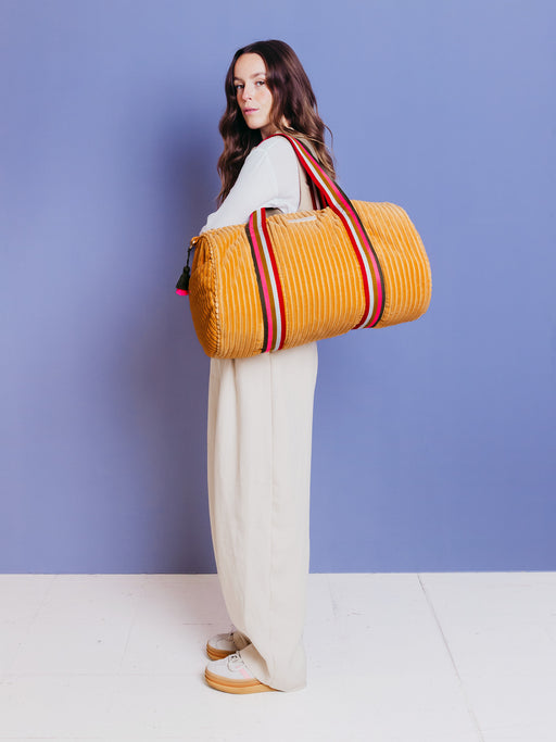 Frau trägt die orangefarbene mara mea Weekender-Tasche True Colors Peach aus Cord an bunten Tragegriffen über der Schulter vor blauem Hintergrund