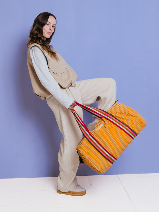 Frau trägt die orangefarbene mara mea Weekender-Tasche True Colors Peach aus Cord an bunten Tragegriffen vor blauem Hintergrund