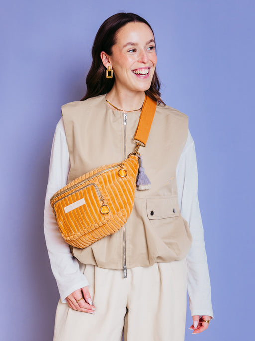 Frau trägt die orangefarbene mara mea Midi Bauchtasche Sunset Date Peach aus Cord quer über der Brust vor blauem Hintergrund