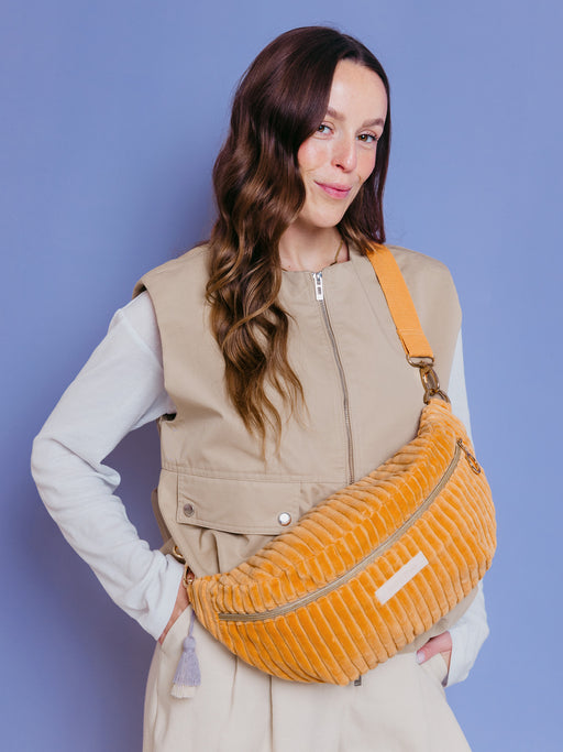 Frau trägt die orangefarbene mara mea Maxi Bauchtasche Soft Serve Peach aus Cord quer über der Brust vor blauem Hintergrund