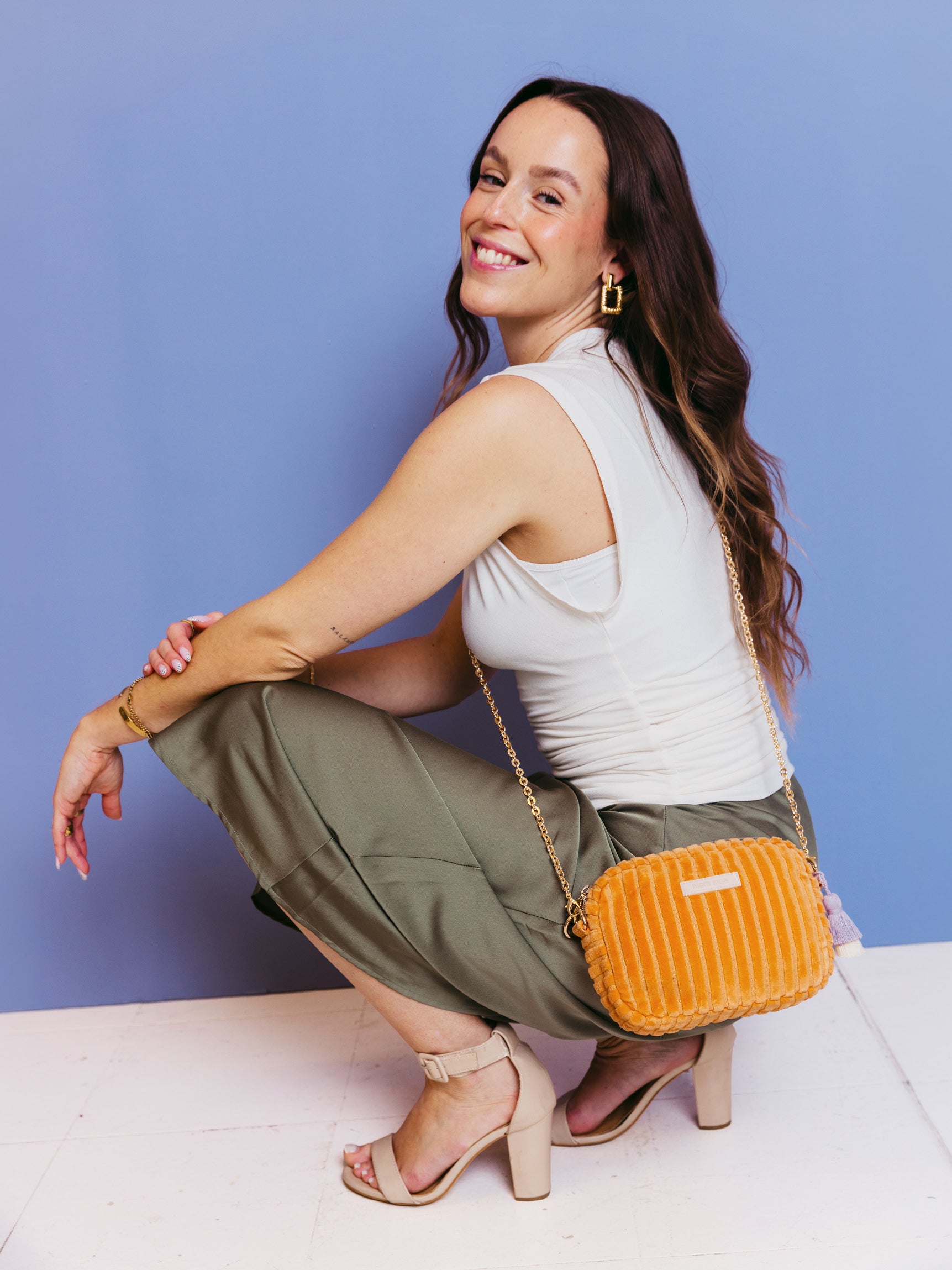 Sitzende Frau trägt die orangefarbene mara mea Umhängetasche Peach Please Peach aus Cord mit goldfarbener Kette vor blauem Hintergrund