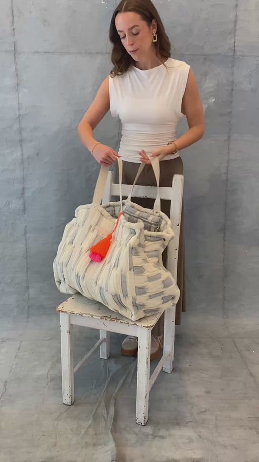 Model zeigt die extra große mara mea Schultertasche daydream dust in Hellblau-Offwhite auf einem Stuhl mit Baumwollgriffen und neon-orange-pinker Quaste